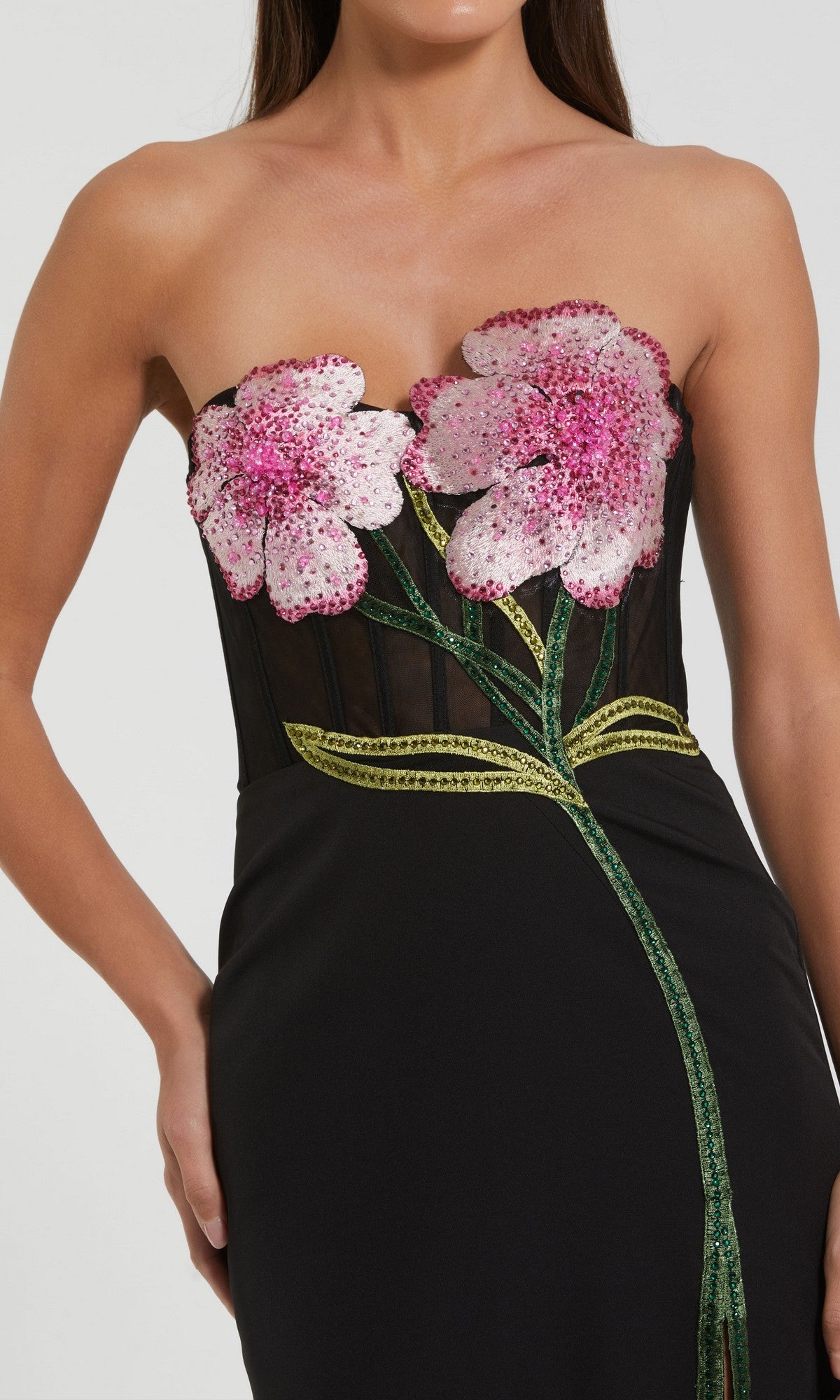 A woman wears the Mac Duggal 20682 Long Formal Dress, a strapless crepe gown featuring pink flower appliqués on the bodice and green stem and leaf embroidery down the skirt—ideal for special occasions.